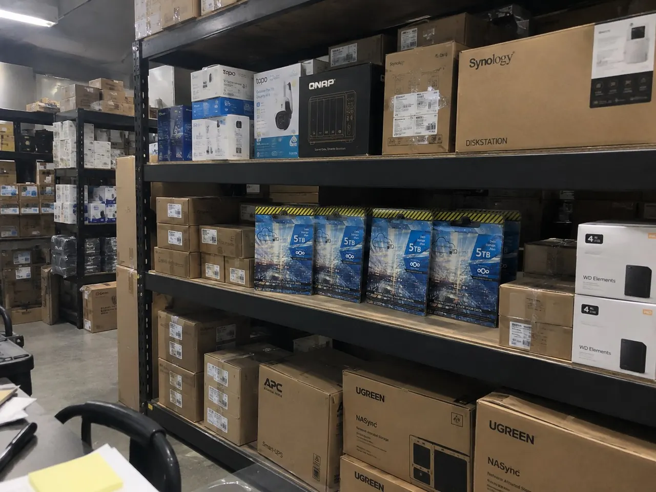 Warehouse shelves stocked with Synology, QNAP, WD Elements, UGREEN NAS, Seagate 5TB, Tapo and APC Smart-UPS storage and networking products